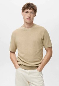 Pullover tricotat cu mânecă scurtă de culoare bej, cu un decolteu rotund, având o suprafață texturată și un tiv côrbă. Purtați cu pantaloni de culoare deschisă.