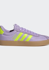 Lavendelfarvet lavtop-sneaker med neon-gule Adidas-striber og logo, gummisål og matchende lavendelfarvede snørebånd på hvid baggrund.