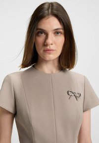 Beige lyhythihainen mekko, jossa on pyöreä kaula-aukko. Mekossa on metallinen rusetti-solki vasemmalla puolella ja teksturoitu kangas.