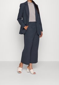 Granatowa, dopasowana marynarka oraz spodnie o szerokich nogawkach w połączeniu z fakturowanym białym i granatowym topem. Beżowe, odkryte mule na średnim obcasie dopełniają outfit.