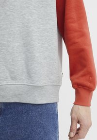 Sudadera gris con una manga roja, que cuenta con una tela texturizada, puños acanalados y una pequeña etiqueta de logo en el dobladillo. Jeans de mezclilla visibles por debajo.