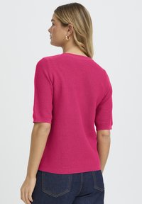 Fuksija pulover s kratkim rukavima, rebraste teksture i okruglog izreza. Nošen s tamnim trapericama, ima pripijeni kroj i suptilan detalj na rubu.