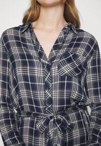 Camicia a quadri navy e bianca con colletto, taschino sul petto e cinturino in vita. Tessuto morbido con una finitura leggermente strutturata.