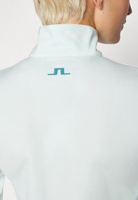 Light blue athletic top with a high collar, featuring a small teal logo on the back. Smooth texture and fitted design.