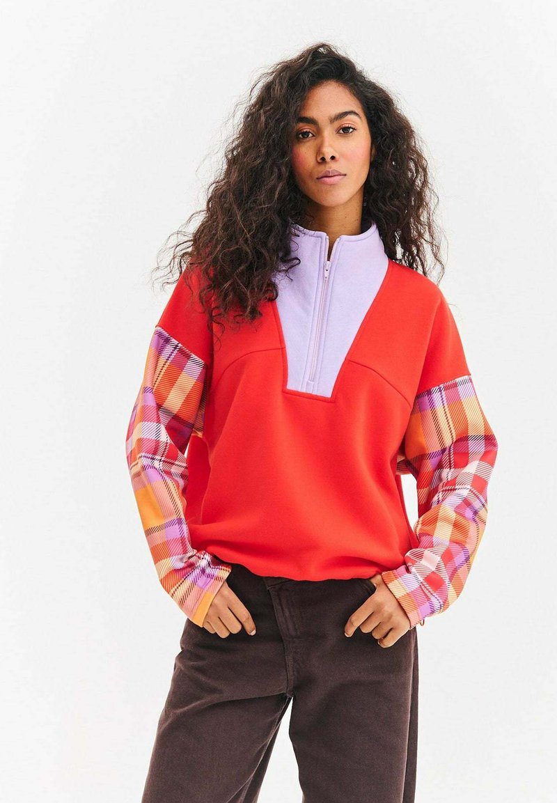 Czerwona bluza z półgolfem na zamek w kolorze fioletowym oraz rękawami w kratę w odcieniach pomarańczu i różu. Gładka tkanina, luźny krój z bocznymi kieszeniami.
