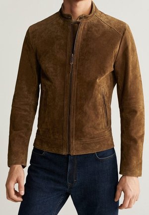 Mies pukeutuneena ruskeaan mokkatakkiseen vetoketjulliseen takkiin, jossa pystykaulus, ja tummansinisiin farkkuihin, seisoo tasaisen vaalean taustan edessä.