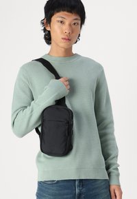 Black fabric crossbody bag with a zippered front pocket, worn over a light green textured sweater. The bag features a shoulder strap.