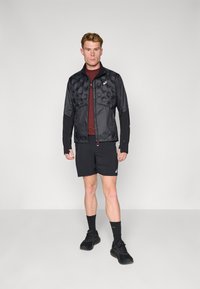 Schwarze Pufferjacke mit hohem Kragen und Reißverschluss, ausgestattet mit einem gesteppten Oberkörper und kontrastierenden Stretchärmeln. Getragen über einem burgunderfarbenen Hemd.