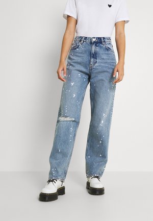Person iført højtaljede blå jeans med hvide malingspletter og en revne på venstre knæ, hvid T-shirt med lille hjerte og hvide snørebåndssko.