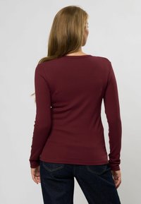 Cămașă burgundy cu mânecă lungă și decolteu rotund, cu un design ajustat, asortată cu blugi de denim închis. Materialul pare moale și elastic.