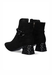 Botines de ante negro hasta el tobillo con punta afilada, que presentan un tacón esculpido negro brillante y un detalle decorativo de cadena negra en el lado.
