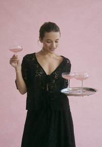 Schwarze Spitzenbluse mit gebundenem Vorderteil, kombiniert mit einem passenden Rock. Hält ein silbernes Tablett mit drei rosa Cocktails. Sanfter rosa Hintergrund.