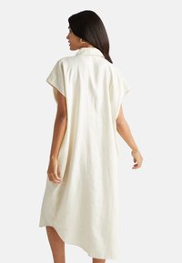 Vestido de lino ligero, color crema, con cuello, mangas cortas y un dobladillo asimétrico, que presenta una silueta relajada y fluida.