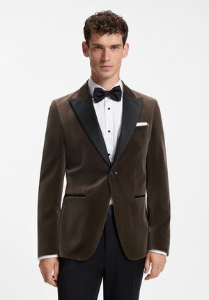 Junger Mann mit lockigem Haar, der eine dunkelbraune Samt-Smokingsjacke, eine schwarze Fliege, ein weißes Hemd und schwarze Hosen vor einem einfarbigen Hintergrund trägt.