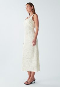 Kleid in Creme mit schwarzen Punkten, ärmelloses Design, knielange Länge, glatte Textur und ergänzt durch schwarze Absatzsandalen.