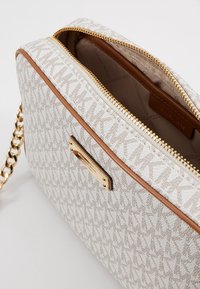 Beżowa torebka typu crossbody Michael Kors w wzór, z złotym zamkiem, brązowymi wykończeniami i złotym łańcuszkiem, częściowo otwarta, aby pokazać podszewkę wewnętrzną.