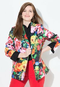 Blommig blazer med svart bas, prydd med livfulla rosor och botaniska mönster. Kombinerad med en svart topp och röda byxor, som visar upp en skräddarsydd design.