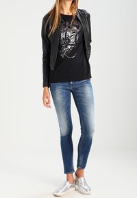 Chaqueta de cuero negra, camiseta gráfica negra, jeans ajustados azules y zapatillas plateadas sin cordones. Texturas suaves y ajuste casual.