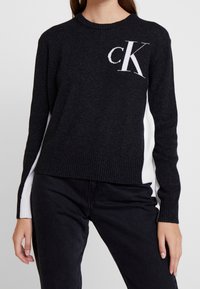 Svart tröja med vita sidpaneler och en vit "CK"-logotyp på bröstet. Ribbad halsringning och muddar, tillverkad av mjukt stickat material.