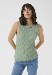 Grøn sansærmet top lavet af tekstureret stof med en bølget halsudskæring og en afslappet pasform. Båret med blå jeans.