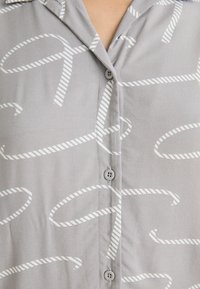 Svetlo siva majica iz mehkega materiala z belim črtastim vzorcem vrvi. Ima zapenjanje s pentljami in klasičen ovratnik.