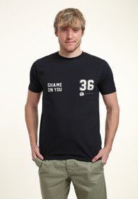 Černé bavlněné tričko s bílým textem "SHAME ON YOU" a číslem "36" na přední straně. Krátké rukávy, design s kulatým výstřihem.