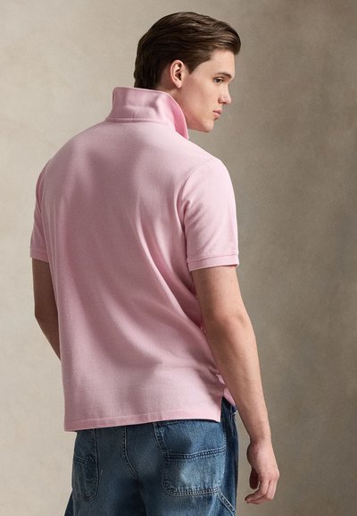 Young man in light pink short-sleeve polo with popped collar, wearing blue denim jeans, standing sideways against neutral background.