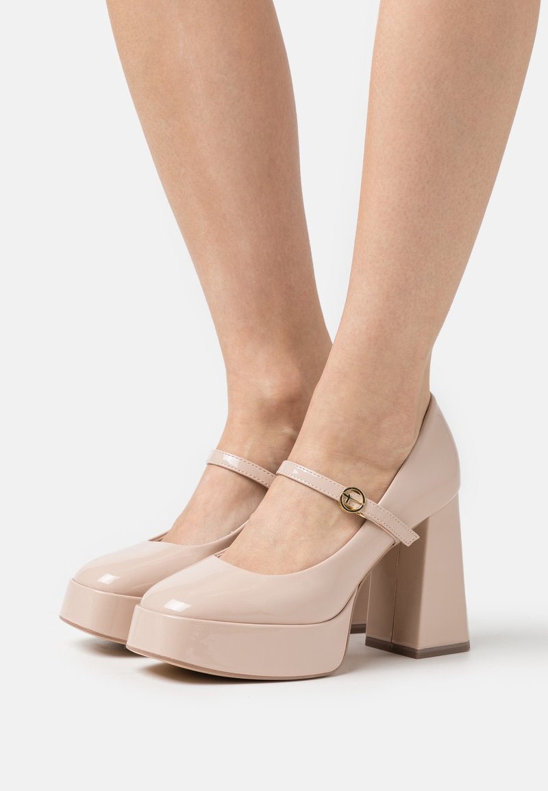 Jambes portant des escarpins Mary Jane à plateforme en cuir verni couleur nude, avec bouts arrondis et brides à boucle dorée sur fond blanc.