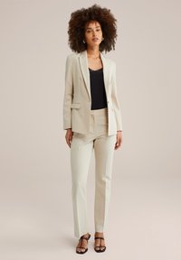 Frau mit lockigem Haar, die einen cremefarbenen Anzug mit maßgeschneiderten Hosen und Blazer über einem schwarzen Spitzenoberteil trägt und beigen Sandalen steht.