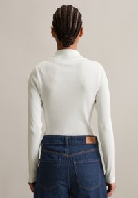 Persoon gezien van achteren met een gevlochten haarbun, in een witte ribgebreide lange mouw top en high-waisted blauwe denim jeans met achterzakken.