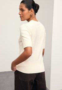 Pull en maille crème à manches courtes, ourlet côtelé et détail torsadé au dos, associé à un pantalon marron foncé. Tissu texturé.