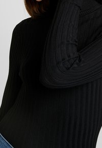 Pull à col roulé noir à côtes avec des manches longues. Présente un tissu texturé et une coupe ajustée, mettant en valeur des lignes verticales sur toute la surface.