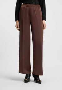Bruine hoge taille wijde broek gecombineerd met zwarte enkellaarsjes met hoge hak en gouden gespsluiting, gedragen door een persoon met een zwart langemouwen shirt.