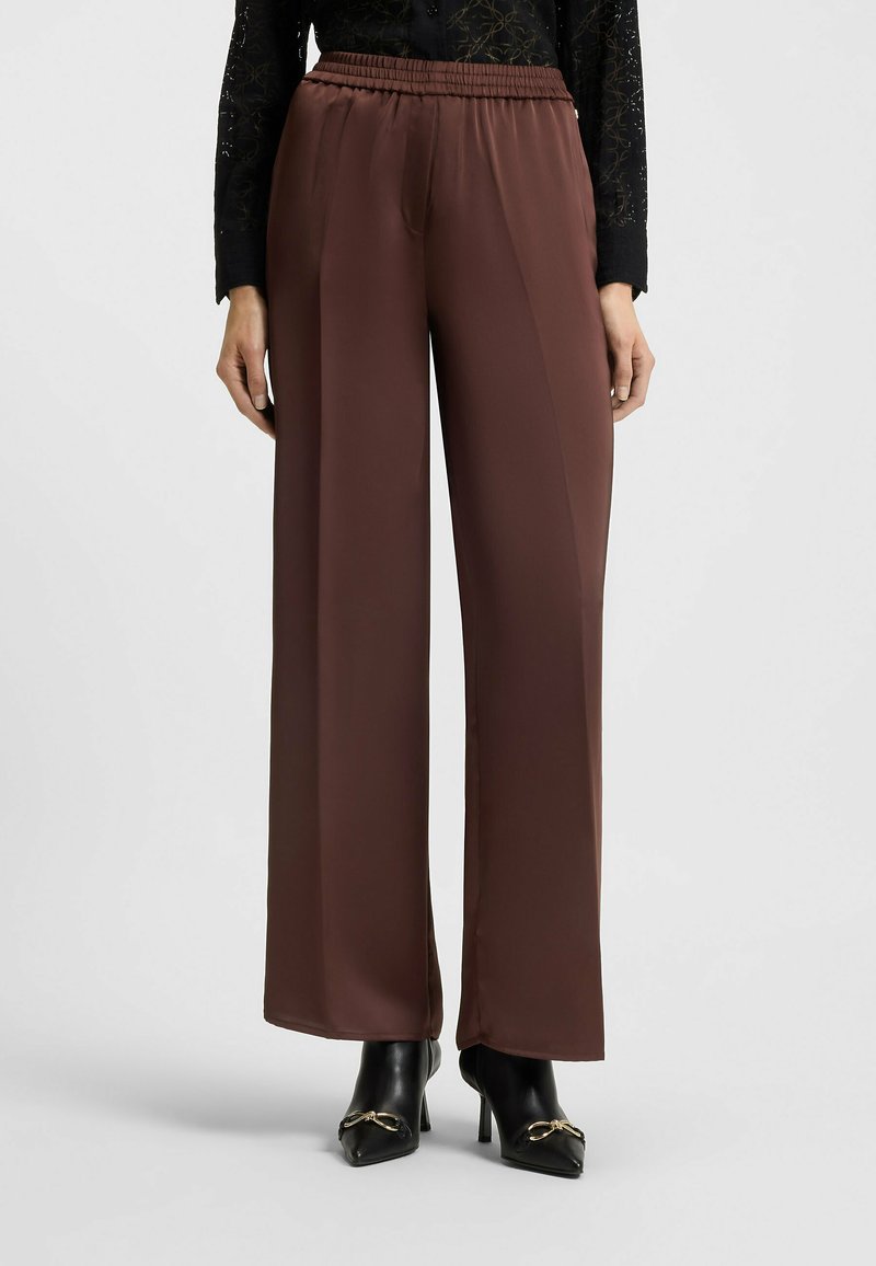 Bruine hoge taille wijde broek gecombineerd met zwarte enkellaarsjes met hoge hak en gouden gespsluiting, gedragen door een persoon met een zwart langemouwen shirt.