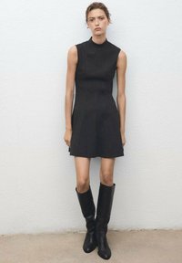 Black sleeveless dress with a high neckline, fitted bodice, and flared skirt. Worn with knee-high black boots. Smooth texture, minimal design.