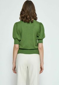 Maglione verde lavorato a maglia con maniche a sbuffo e orlo a coste, abbinato a pantaloni chiari. Texture morbida e design aderente.