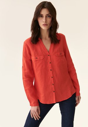 Rød linned skjorte med lange ærmer, V-udskæring, to brystlommer og knaplukning. Glat tekstur og afslappet pasform. Båret med mørke jeans.