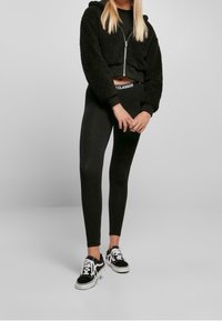 Czarna krótka kurtka polarowa z kapturem, w zestawie z czarnymi spodniami w stylu legginsów. Obuwie to czarne sneakersy z białymi akcentami.
