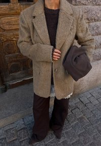 Personne portant un blazer beige duveteux sur un pull sombre et un chemisier en dentelle, tenant un chapeau plié marron foncé dans une rue pavée.