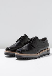 Chaussures brogue en cuir noir avec des détails perforés. Elles disposent d'une semelle en caoutchouc texturé léger avec un intercalaire en bois pour le contraste.