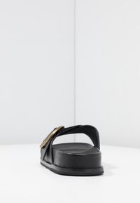 Černé kožené páskové sandály s širokým popruhem, zdobené zlatým kruhovým akcentem. Plochá podrážka a minimalistický design, který zdůrazňuje texturu a siluetu.