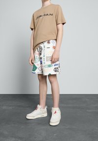 Hnědá tričko s potiskem "JORDAN", bílé šortky s barevnými logy a krémové tenisky s červenými akcenty a texturovanými detaily.