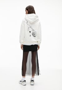 Haut à capuche blanc avec un graphique de cœur en porte-clés au dos. Associé à une jupe noire avec un panneau transparent. Baskets blanches avec accents noirs.