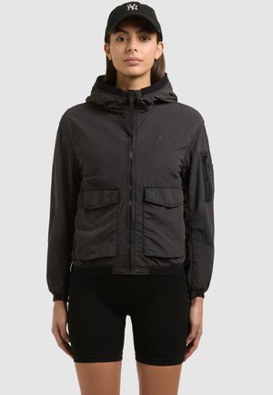 Nő fekete cipzáras kapucnis dzsekiben első zsebekkel, fekete rövidnadrágban és fekete sapkában fehér logóval, egyszínű háttér előtt állva.