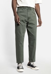 Olive green cotton trousers with a straight cut, featuring front pockets, a zipper fly, and worn with black canvas sneakers.