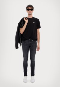 T-shirt nera con logo, giacca di pelle nera lasciata sulla spalla, jeans skinny grigi e sneaker bianche. Occhiali da sole eleganti completano il look.