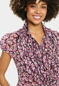 Gėlėta bluzonė su rožinėmis, juodomis ir baltomis spalvomis, pagaminta iš permatomo audinio. Rūbui būdinga volangų dekoracija prie apykaklės ir priekyje, taip pat pūstinės rankovės.