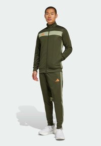 Green sports tracksuit made of smooth fabric, featuring a zip-up jacket with a light green panel and orange logo, and matching tapered pants with stripes.