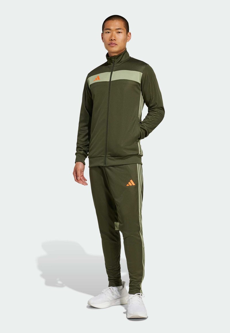 Green sports tracksuit made of smooth fabric, featuring a zip-up jacket with a light green panel and orange logo, and matching tapered pants with stripes.