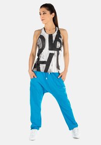 Zwarte en witte tanktop met abstract ontwerp; helder blauwe harem broek; model draagt witte sneakers en staat tegen een effen achtergrond.